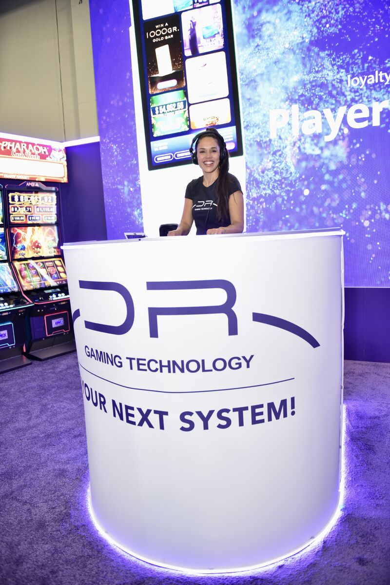 Anfitriona en el mostrador de DR Gaming Technology con el lema Your Next System y máquinas de juego al fondo