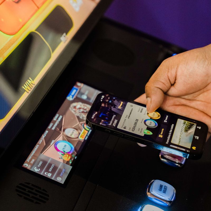 Un jugador acerca su teléfono móvil a una máquina tragamonedas mostrando la app de billetera con saldo y promociones